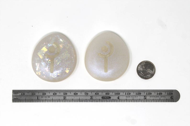 May include: Two white oval-shaped stones with a silver design of a wand and crescent moon. The stones are on a white surface with a ruler for scale.