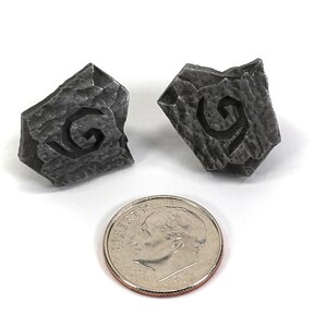 Puede incluir: Dos dados peque&ntilde;os de metal gris con una letra G estilizada tallada en cada cara. Los dados son aproximadamente del tama&ntilde;o de una moneda de diez c&eacute;ntimos de euro.