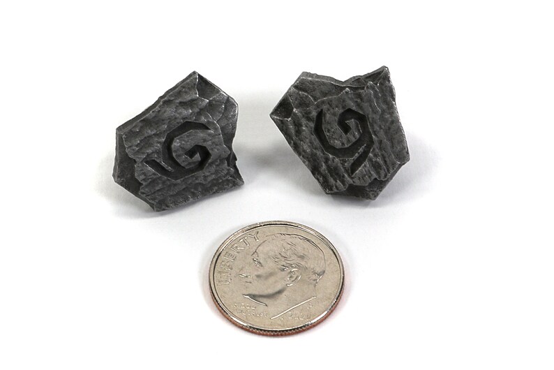 Puede incluir: Dos dados peque&ntilde;os de metal gris con una letra G estilizada tallada en cada cara. Los dados son aproximadamente del tama&ntilde;o de una moneda de diez c&eacute;ntimos de euro.