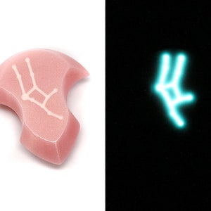 May include: A pink ceramic pendant with a white outline of the Gemini constellation. The pendant glows a light blue in the dark.