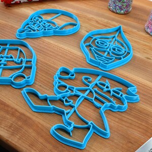 Puede incluir: Cuatro cortadores de galletas de pl&aacute;stico azul sobre una superficie de madera. Los cortadores tienen forma de coche, persona, cara y sombrero.