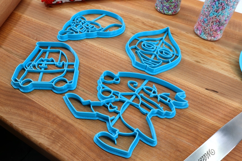 Puede incluir: Cuatro cortadores de galletas de pl&aacute;stico azul sobre una superficie de madera. Los cortadores tienen forma de coche, persona, cara y sombrero.