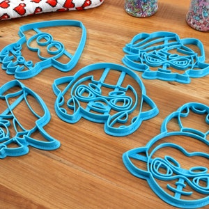 May include: Five blue plastic cookie cutters shaped like cartoon characters with large eyes and goggles. The characters are all wearing hats.