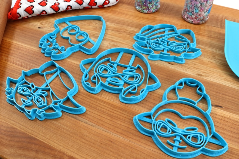 May include: Five blue plastic cookie cutters shaped like cartoon characters with large eyes and goggles. The characters are all wearing hats.