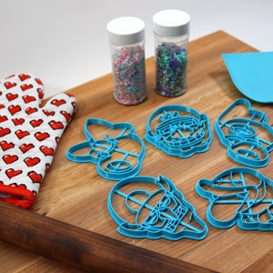 May include: A set of blue plastic cookie cutters shaped like cartoon characters, a wooden rolling pin, a red oven mitt with white hearts, and two jars of sprinkles on a wooden cutting board.