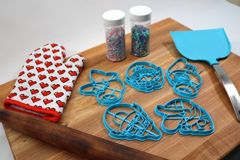 May include: A set of blue plastic cookie cutters shaped like cartoon characters, a wooden rolling pin, a red oven mitt with white hearts, and two jars of sprinkles on a wooden cutting board.