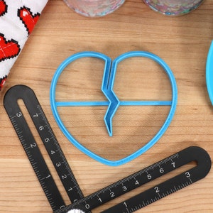 May include: A blue plastic cookie cutter shaped like a broken heart. The cutter is on a wooden surface with a ruler and a blue silicone spatula.