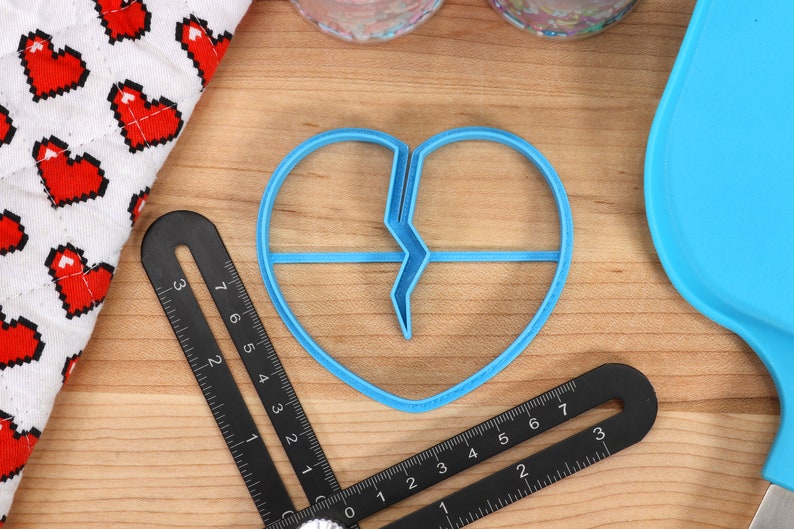May include: A blue plastic cookie cutter shaped like a broken heart. The cutter is on a wooden surface with a ruler and a blue silicone spatula.