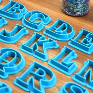 May include: A set of blue plastic alphabet cookie cutters on a wooden surface. The cutters are arranged in alphabetical order.
