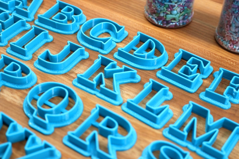 May include: A set of blue plastic alphabet cookie cutters on a wooden surface. The cutters are arranged in alphabetical order.
