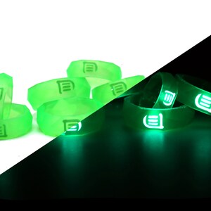 May include: A set of green glow-in-the-dark silicone rings with a white logo. The rings are stacked on a white surface and glow green in the dark.