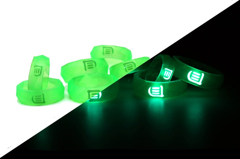 May include: A set of green glow-in-the-dark silicone rings with a white logo. The rings are stacked on a white surface and glow green in the dark.