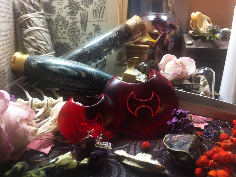 May include: Two red, round, glass pendants with a black symbol in the center. The pendants are on a dark surface with dried flowers and other decorative items.