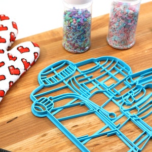 May include: Blue plastic cookie cutters shaped like a coffee cup and a coffee bean on a wooden surface. There are two jars of sprinkles in the background, one with pink and white sprinkles and the other with multi-colored sprinkles.