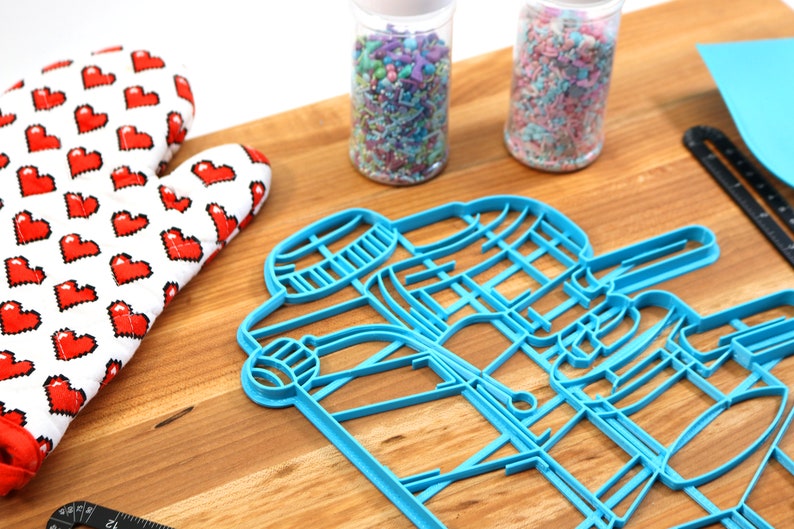 May include: Blue plastic cookie cutters shaped like a coffee cup and a coffee bean on a wooden surface. There are two jars of sprinkles in the background, one with pink and white sprinkles and the other with multi-colored sprinkles.