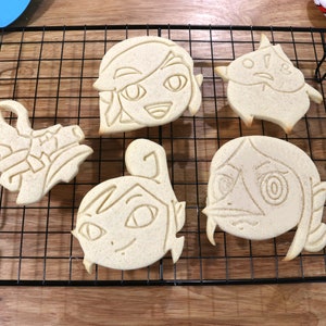 May include: Five unbaked sugar cookies on a black wire cooling rack. The cookies are shaped like cartoon characters with detailed facial features.