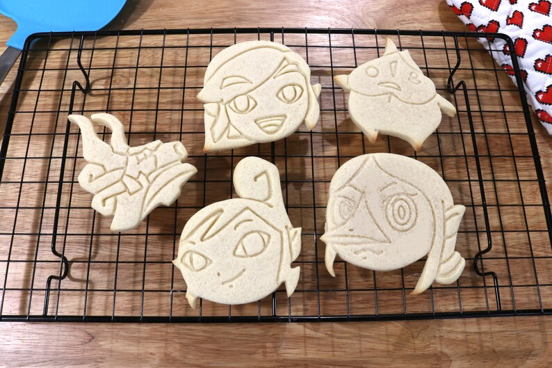 May include: Five unbaked sugar cookies on a black wire cooling rack. The cookies are shaped like cartoon characters with detailed facial features.