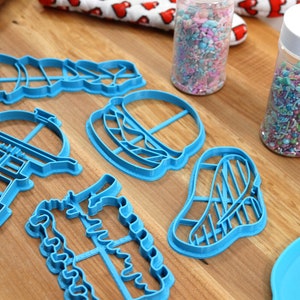 K&ouml;nnte beinhalten: Ein Set mit sechs blauen Plastik-Ausstechern in Form eines Grills, eines Hamburgers, eines Steaks, eines Weihnachtsbaums, eines Kaktus und eines Leuchtturms. Die Ausstecher liegen auf einer Holzoberfl&auml;che mit zwei Gl&auml;sern Streusel.