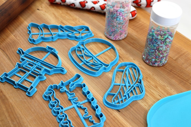 K&ouml;nnte beinhalten: Ein Set mit sechs blauen Plastik-Ausstechern in Form eines Grills, eines Hamburgers, eines Steaks, eines Weihnachtsbaums, eines Kaktus und eines Leuchtturms. Die Ausstecher liegen auf einer Holzoberfl&auml;che mit zwei Gl&auml;sern Streusel.