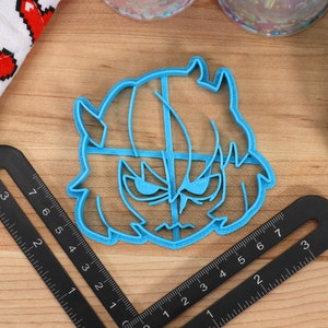 May include: A blue plastic cookie cutter shaped like a cartoon character with horns, a frown, and a hairstyle. The cookie cutter is on a wooden surface with a ruler.