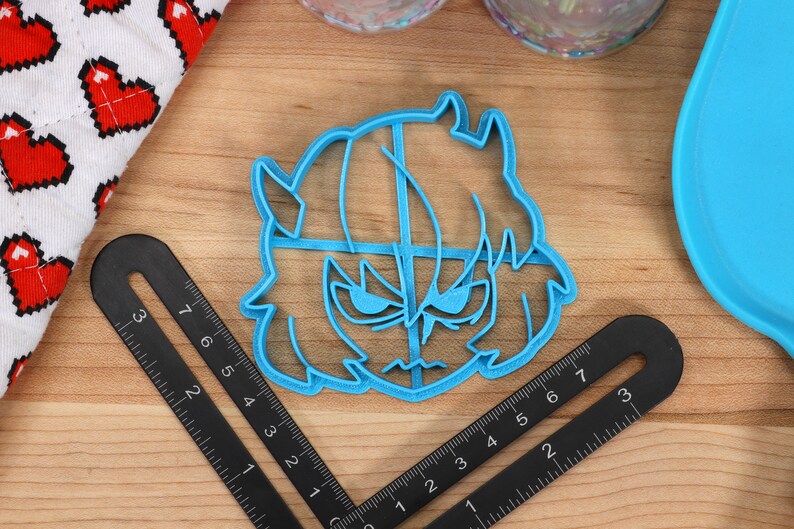 May include: A blue plastic cookie cutter shaped like a cartoon character with horns, a frown, and a hairstyle. The cookie cutter is on a wooden surface with a ruler.