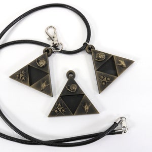 May include: Three gold-colored triangular pendants with a black cord necklace. Each pendant features a different design with a stylized bird, star, and other symbols.