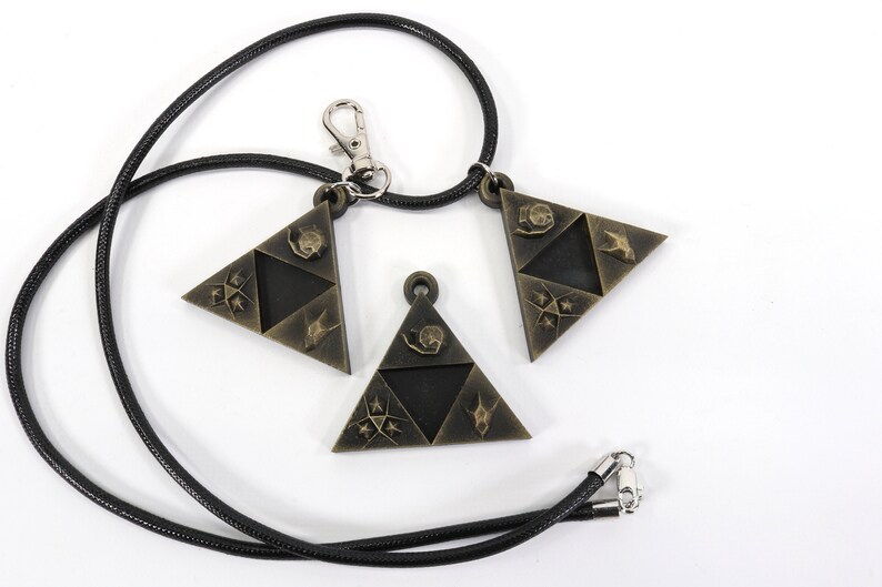 May include: Three gold-colored triangular pendants with a black cord necklace. Each pendant features a different design with a stylized bird, star, and other symbols.