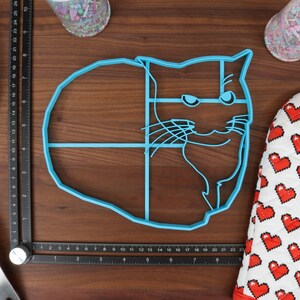 May include: A blue plastic cat-shaped cookie cutter. The cutter is divided into sections with lines. The cutter is on a wooden surface.