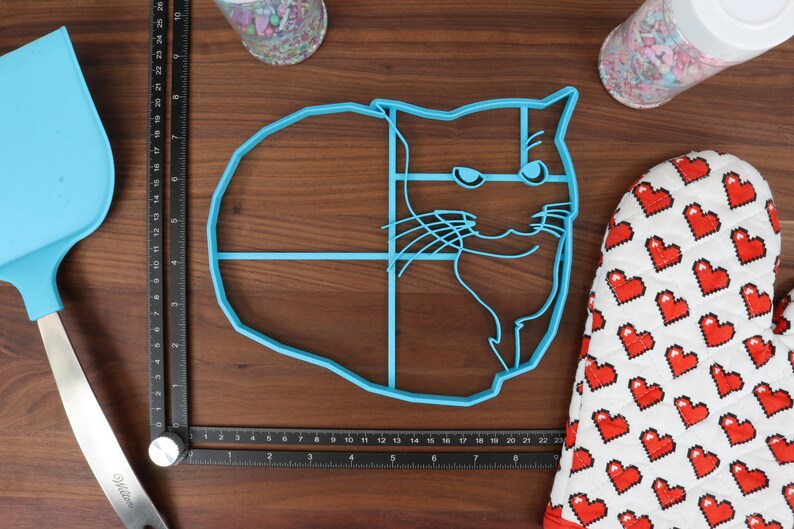 May include: A blue plastic cat-shaped cookie cutter. The cutter is divided into sections with lines. The cutter is on a wooden surface.