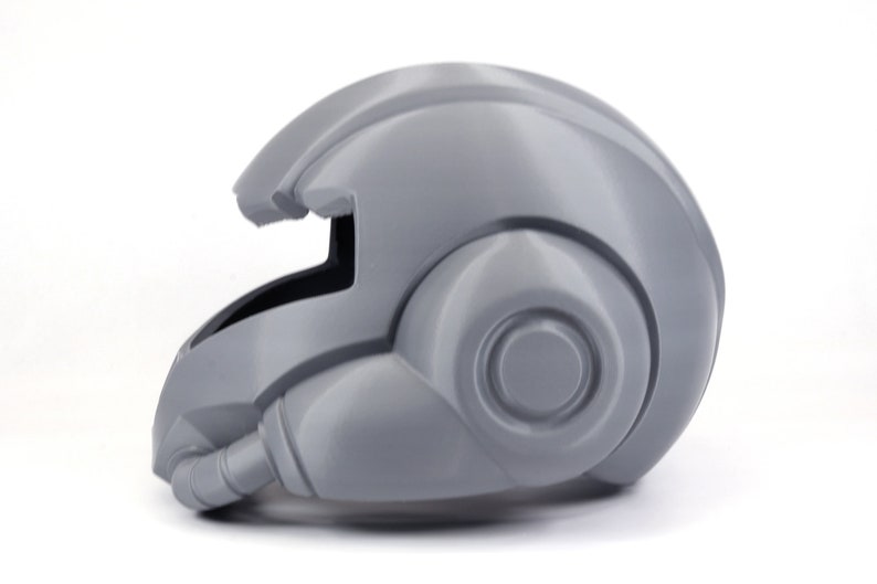 May include: A gray 3D printed helmet with a futuristic design. The helmet has a visor and a large, round earpiece on the right side.