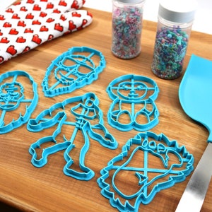 May include: A set of six blue plastic cookie cutters shaped like cartoon characters. The cutters are on a wooden surface with a rolling pin, a spatula, and two jars of sprinkles.
