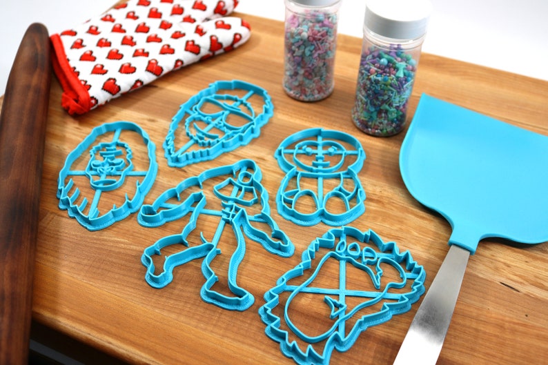 May include: A set of six blue plastic cookie cutters shaped like cartoon characters. The cutters are on a wooden surface with a rolling pin, a spatula, and two jars of sprinkles.