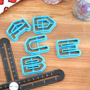 May include: A set of five blue plastic cookie cutters shaped like the letters A, C, E, and two Bs. The cutters are on a wooden surface with a black ruler and a white and red fabric with hearts in the background.