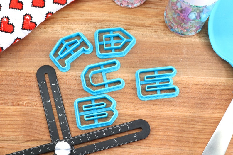 May include: A set of five blue plastic cookie cutters shaped like the letters A, C, E, and two Bs. The cutters are on a wooden surface with a black ruler and a white and red fabric with hearts in the background.