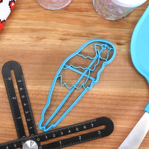May include: A light blue bird-shaped cookie cutter, a blue spatula, and two black angle finders are arranged on a wooden surface. A white oven mitt with red pixelated hearts is in the upper left corner. Two clear jars are in the background.