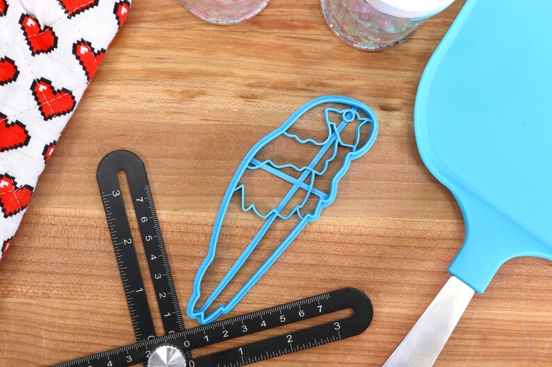 May include: A light blue bird-shaped cookie cutter, a blue spatula, and two black angle finders are arranged on a wooden surface. A white oven mitt with red pixelated hearts is in the upper left corner. Two clear jars are in the background.