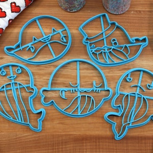 Peut inclure: Cinq emportes-pi&egrave;ces en plastique bleu en forme de baleines avec diff&eacute;rentes expressions faciales. Les emportes-pi&egrave;ces sont sur une surface en bois.