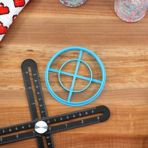 May include: A collection of baking tools on a wooden surface. Includes a blue circle-shaped cookie cutter, a black multi-angle measuring tool, a heart-patterned kitchen towel, and a blue spatula. The tools are for baking.