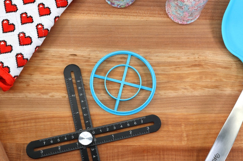 May include: A collection of baking tools on a wooden surface. Includes a blue circle-shaped cookie cutter, a black multi-angle measuring tool, a heart-patterned kitchen towel, and a blue spatula. The tools are for baking.