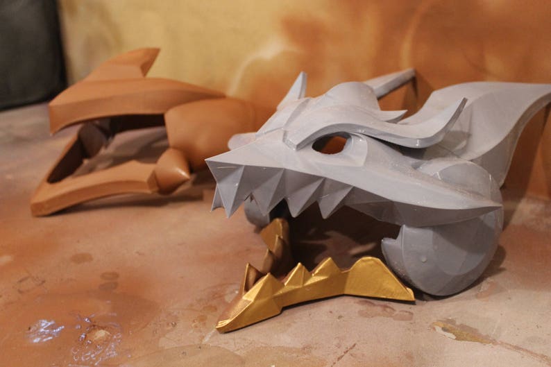 May include: A gray and gold dragon head costume piece with sharp teeth and a textured surface.