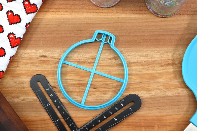 Op de afbeelding: Een blauwe plastic koekjesvormer in de vorm van een kerstbal met een kruisvormig ontwerp erin. De vormer ligt op een houten oppervlak.