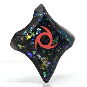 Pu&ograve; includere: Un ciondolo in resina nero a forma di diamante con un simbolo rosso a spirale al centro. Il ciondolo &egrave; ricoperto di glitter iridescente.