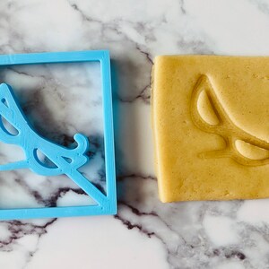 May include: A blue plastic cookie cutter in the shape of a square with a pair of sunglasses inside. The sunglasses are crossed out with a diagonal line. A cookie with the same design is shown next to the cutter.