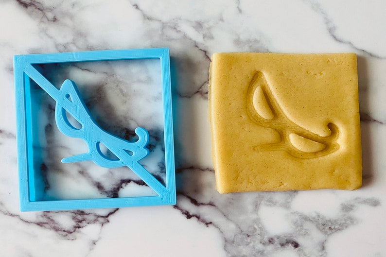 May include: A blue plastic cookie cutter in the shape of a square with a pair of sunglasses inside. The sunglasses are crossed out with a diagonal line. A cookie with the same design is shown next to the cutter.