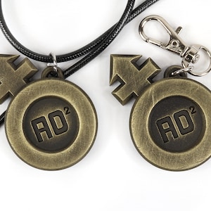 May include: Two gold-toned metal pendants with a black cord and a silver key chain. Each pendant features a stylized arrow symbol and a circular design with the text "AD&sup2;" in black.