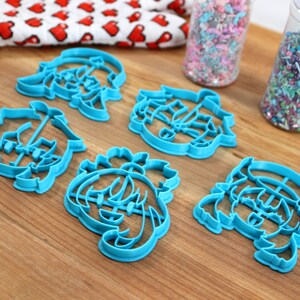 May include: Five blue plastic cookie cutters shaped like cartoon characters with different hairstyles. The cutters are on a wooden surface.
