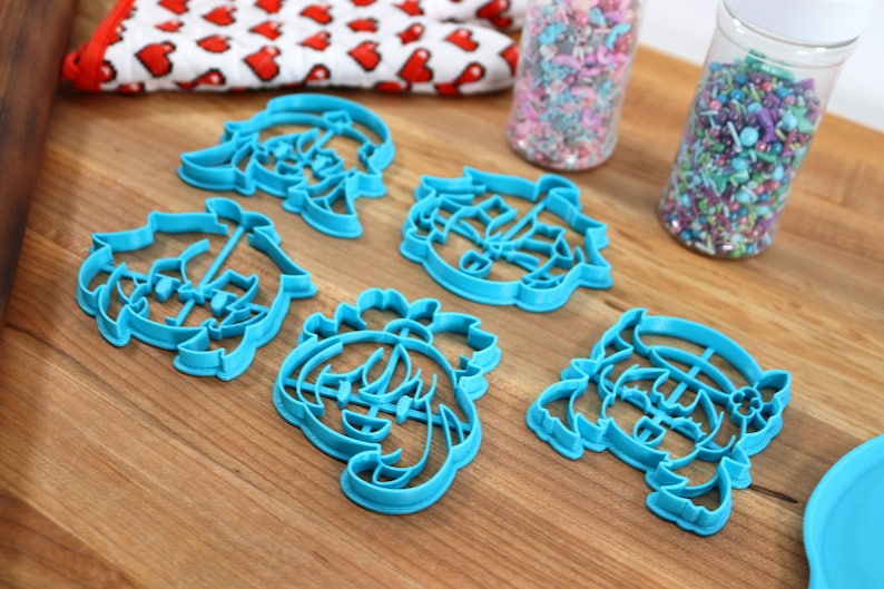 May include: Five blue plastic cookie cutters shaped like cartoon characters with different hairstyles. The cutters are on a wooden surface.