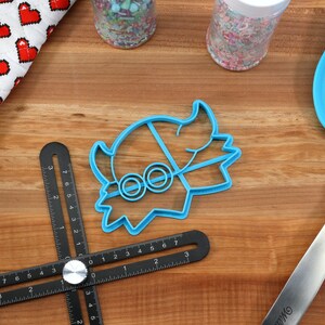 May include: A blue cookie cutter in the shape of a cartoon character with round eyes, a blue spatula, and a black measuring tool are on a wooden surface. A red and white oven mitt with heart shapes and jars of sprinkles are also present.