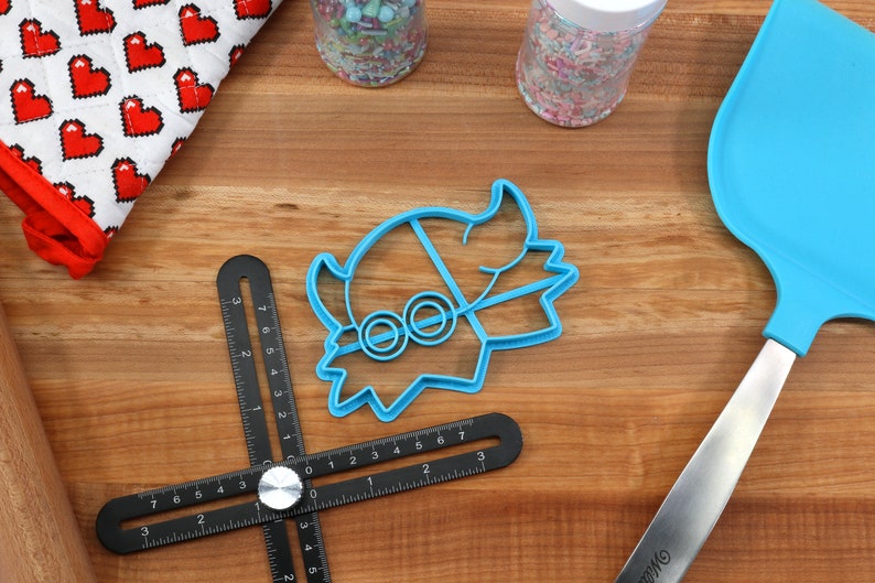 May include: A blue cookie cutter in the shape of a cartoon character with round eyes, a blue spatula, and a black measuring tool are on a wooden surface. A red and white oven mitt with heart shapes and jars of sprinkles are also present.