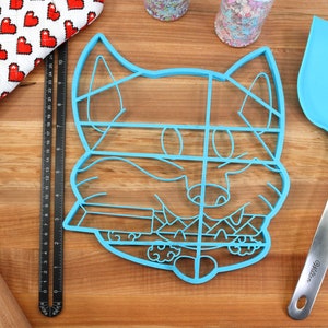 May include: A blue plastic cookie cutter shaped like a cartoon fox's face. The cutter is divided into sections for decorating cookies.
